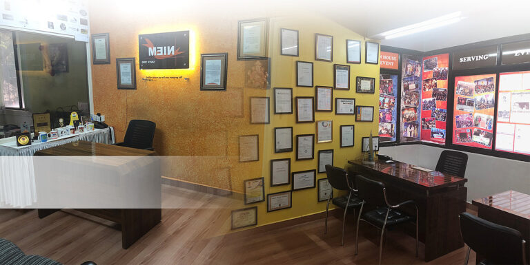 Office space decorated with framed certificates and accolades on the walls, a meeting area with a table and chairs to the left, and a showcase of colorful posters or achievements to the right, under a warm and inviting lighting atmosphere.